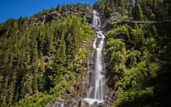 Wasserfall in bergiger Landschaft mit grünen Bäumen umgeben von Wald unter blauem Himmel.