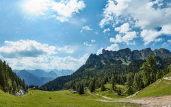 Panorama einer alpinen Landschaft mit Wiesen und Bergen unter blauem Himmel.