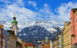 Stadtansicht mit bunten Gebäuden und schneebedeckten Bergen im Hintergrund.