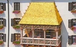 Balkon mit goldenem Dach und bunten Blumen an Fenstern einer weißen Fassade.