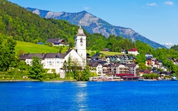 Bergdorf am See mit Kirche im Vordergrund, umgeben von grünen Hügeln und bewaldeten Bergen.