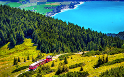 Roter Zug fährt durch Waldlandschaft neben blauem See.