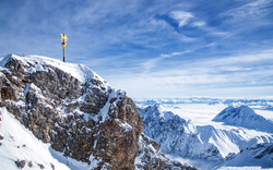 Schneebedeckter Berggipfel mit einem markanten goldenen Gipfelkreuz.