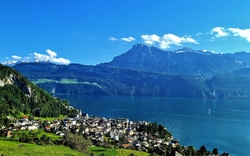 Blick auf ein Dorf am See vor einer Berglandschaft bei klarem Himmel.