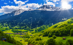 Blick auf schneebedecktes Gebirge und grünes Tal unter blauem Himmel mit Sonne.
