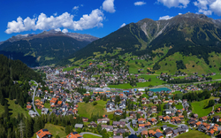 Luftaufnahme eines Dorfes in den Alpen mit grünen Wiesen und Bergen im Hintergrund.