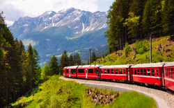 Roter Zug fährt durch grüne Berglandschaft mit schneebedeckten Bergen im Hintergrund.