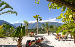 Palmen und Liegestühle an einem Strand mit Blick auf Berge und blauen Himmel.