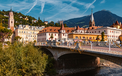 Blick auf eine Brücke über einen Fluss vor malerischen Gebäuden und Bergkulisse.