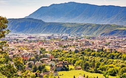 Stadtansicht mit grünen Bäumen und Bergen im Hintergrund bei sonnigem Wetter.