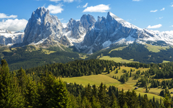 Berglandschaft mit bewaldeten Hügeln und schneebedeckten Gipfeln unter blauem Himmel.