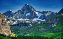 Berglandschaft mit schneebedecktem Gipfel und grünen Hügeln unter blauem Himmel.