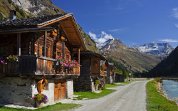 Holzhäuser an einem Bergweg mit blühenden Blumen und schneebedeckten Gipfeln.