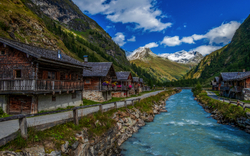 Bergdorf mit Holzhäusern und Fluss vor schneebedeckten Alpengipfeln