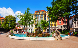 Springbrunnen auf einem Stadtplatz mit bunten Gebäuden im Hintergrund.