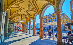 Bogengang mit Säulen und Blick auf einen sonnigen Platz, Menschen sitzen an Tischen.