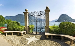Metalltor mit Seeblick, flankiert von grünen Büschen und Blumenbeeten.