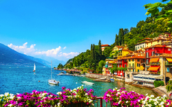 Bunte Häuser am Seeufer mit Blumen im Vordergrund und Segelbooten im Wasser.