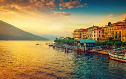 Häuser an einem Seeufer bei Sonnenuntergang mit Boot und Bergen im Hintergrund.