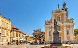 Platz mit Statue, barocker Kirche und Gebäuden unter blauem Himmel.