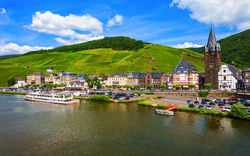 Blick auf ein malerisches Dorf am Fluss mit grünen Hügeln im Hintergrund.