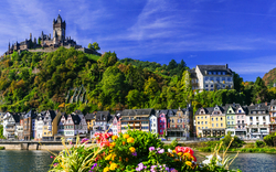 Blick auf historische Burg und bunte Häuser an einem Fluss in malerischer Landschaft.