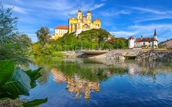 Barockes Schloss auf Hügel spiegelt sich im ruhigen Fluss unter blauem Himmel.