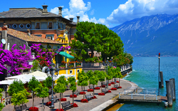 Promenade am See mit Bäumen, Sitzbereichen und Bergen im Hintergrund.
