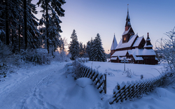 Winterlandschaft mit verschneiter Holzkapelle und Tannenbäumen im Hintergrund.