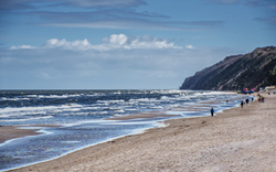 Strand mit Wellen und Menschen am Ufer, bewölkter Himmel im Hintergrund.