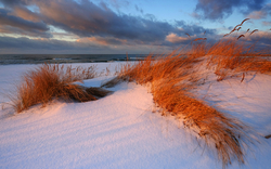 Schneebedeckte Dünen am Meer bei Sonnenaufgang mit Wolken am Himmel.