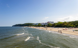 Strand mit Promenade, Menschen spazieren, Gebäude und grüner Hügel im Hintergrund.