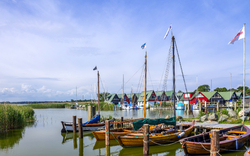 Holzboote auf einem ruhigen See, umgeben von Schilf und kleinen farbigen Gebäuden am Ufer.