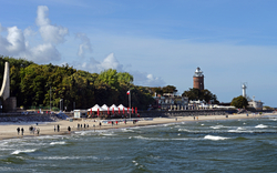Küstenszene mit Strand, Menschen, Leuchtturm und Wolken am blauen Himmel.