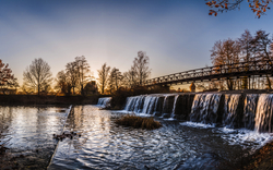 Fluss mit Wasserfall und Brücke in herbstlicher Landschaft bei Sonnenuntergang.