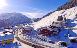 Winterliche Panoramaaufnahme eines verschneiten Bergdorfs mit Alpen im Hintergrund.