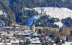 Verschneite Winterlandschaft mit Dorf, Kirche und Skisprungschanzen im Hintergrund.