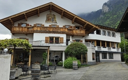 Weißes Haus mit hellem, hölzernem Dach und Balkon, davor eine Terrasse mit Bestuhlung, im Hintergrund eine bergige Landschaft die in die Wolken ragt