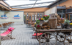Hof der Erlebnissennerei mit rustikalem Wagen und Blumenschmuck, Holzfassade im Hintergrund.