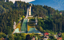 Skisprungschanze umgeben von Wald und Bergen mit umliegenden Gebäuden.