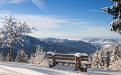 Verschneite Berglandschaft mit Bank im Vordergrund und blauen Himmel.