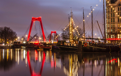 Abendliche Szenerie mit beleuchteter Brücke und Segelbooten im Hafen.