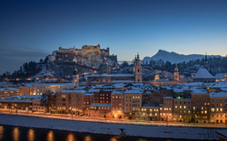 Verschneite Stadt und Burg bei Abenddämmerung, beleuchtet entlang eines Flusses.