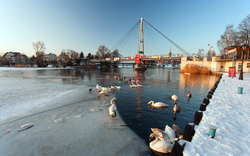 Verschneiter Fluss mit Schwänen und Fußgängerbrücke im Hintergrund.