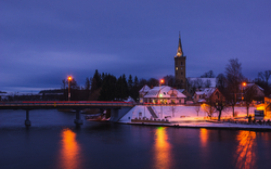 Verschneite Abendlandschaft mit beleuchteter Kirche und Brücke über den Fluss.