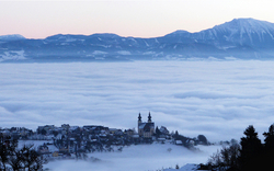 Verschneite Landschaft mit nebelverhangener Stadt und Bergen im Hintergrund.