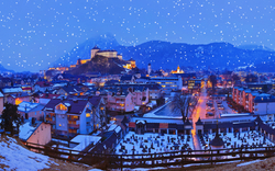 Stadtlandschaft im Winter mit Schneefall und Burg im Hintergrund bei Abenddämmerung.