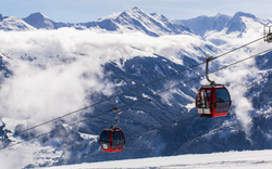 Zwei rote Gondeln vor verschneiten Alpen und bewölktem Himmel.