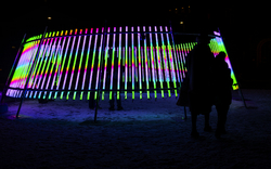 Leuchtinstallation mit bunten vertikalen Lichtstäben und zwei silhouettierten Personen.