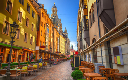 Städtische Gasse mit Restaurants und Kirchturm im Hintergrund bei sonnigem Wetter.